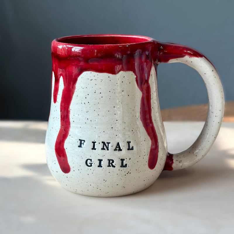A speckled off-white ceramic mug with red dripping glaze and the words 'FINAL GIRL' printed in black on the front, shown on a tabletop against a dark gray background.