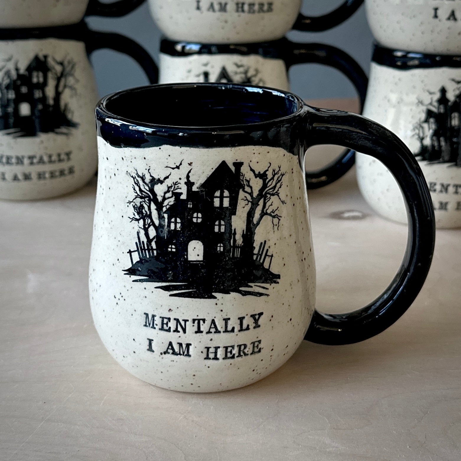 A ceramic mug with a speckled off-white body, black handle, and black interior, printed with a haunted house illustration and the words 'MENTALLY I AM HERE.' Other matching mugs are blurred in the background on a light tabletop.