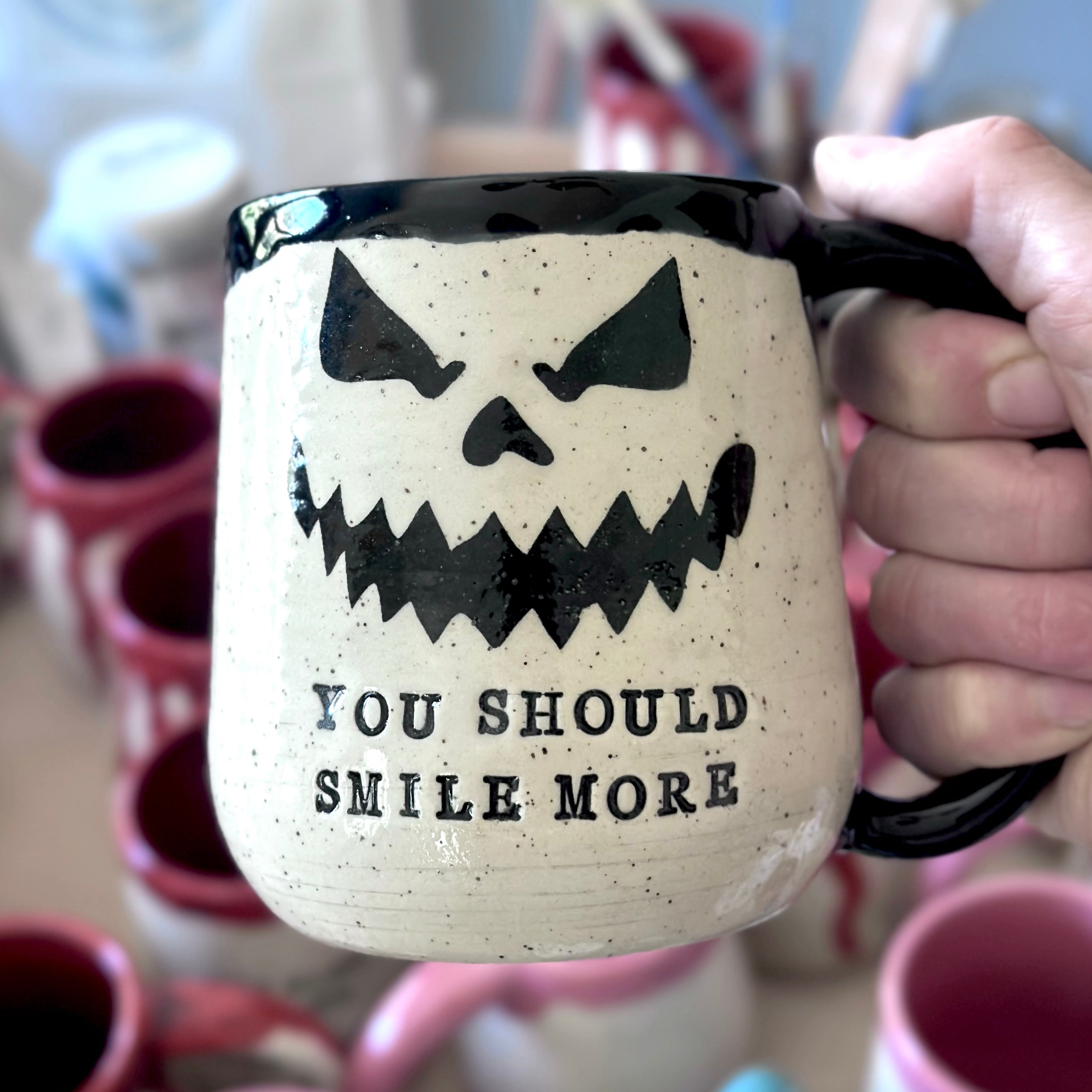 A hand holds a speckled off-white ceramic mug with a black handle and black rim. The mug features a black spooky face graphic and the text 'YOU SHOULD SMILE MORE'.